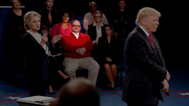 Ken Bone sits in the audience at presidential debate. He is wearing a red sweater, a white shirt and glasses.