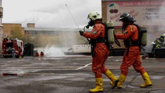 Two people wear hazmat suits with helmets and rubber boots. They have air tanks on their backs.