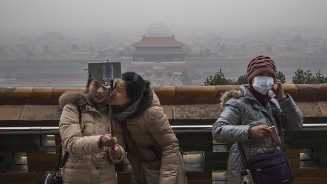 A Chinese woman wears a pollution mask as two others take a selfie