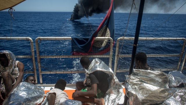 Refugees watch from an aid ship as a migrant boat burns in the Mediterranean