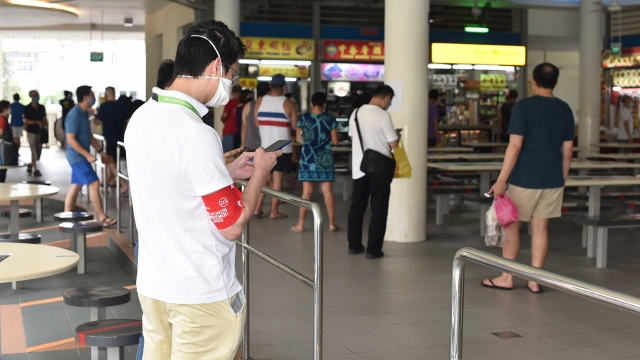 A safe-distancing enforcement officer wearing a red armband checks his phone at a food court in Singapore