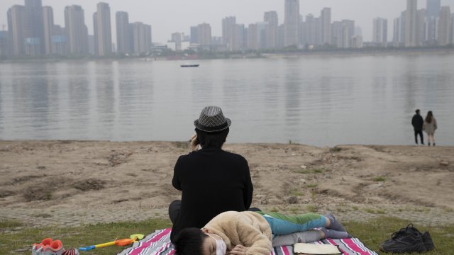 Child wearing a mask lays outside on a blanket along the banks of a Wuhan river