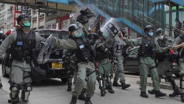 Riot police cover themselves with shields as hundreds of protesters march along a downtown street