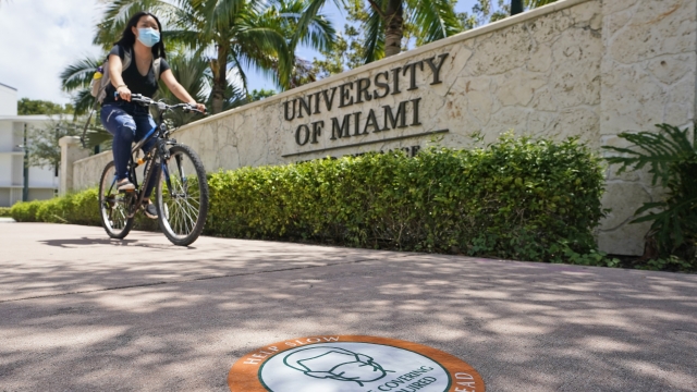 A cyclist, wearing a mask to prevent the spread of coronavirus, rides by an entrance to the University of Miami.