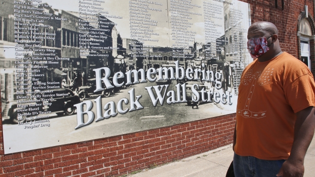 A man stands in front of a mural listing the names of businesses destroyed during the 1921 Tulsa Race Massacre.