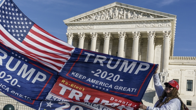 A Trump supporter unfurls a patchwork flag of the American flag and three Trump banners in front of the Supreme Court
