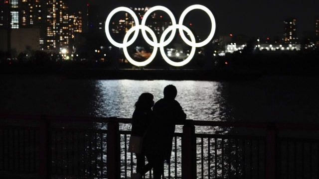 A man and a woman stand with the backdrop of the Olympic rings floating in the water.