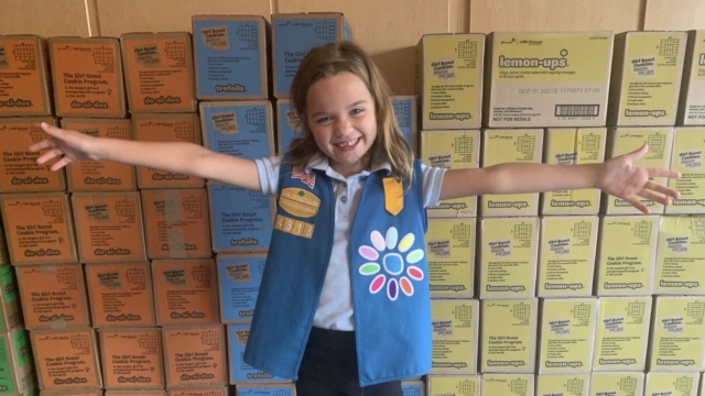 Girl scout poses by cookie boxes.