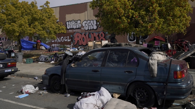 A person stands in front of a tent behind a car filled with debris at a homeless encampment