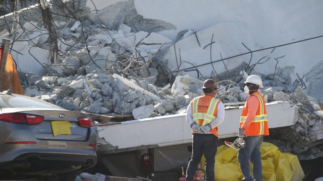 Workers stand next to a crushed car and yellow tarp under a section of a collapsed pedestrian bridge.