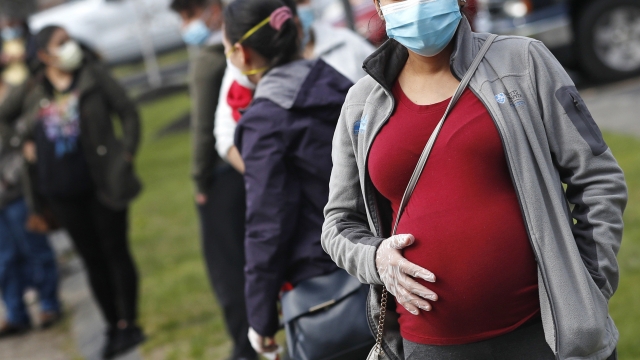 A pregnant woman wearing a face mask and gloves holds her belly as she waits in line for groceries