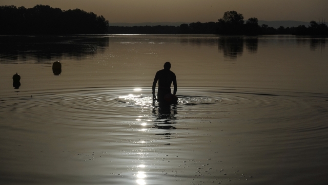 A man walks in the water as the sun rises above the lake.