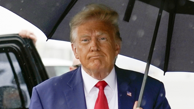 Former President Donald Trump walks to speak with reporters while holding an umbrella.