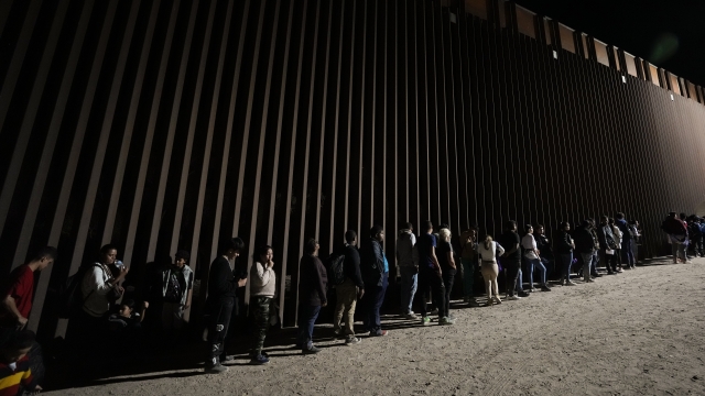 People line up against a border wall as they wait to apply for asylum after crossing the border from Mexico