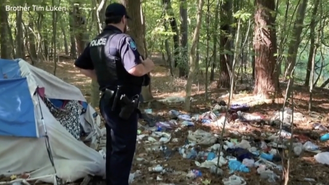 A police officer looks at trash left by homeless people in a wooded area.