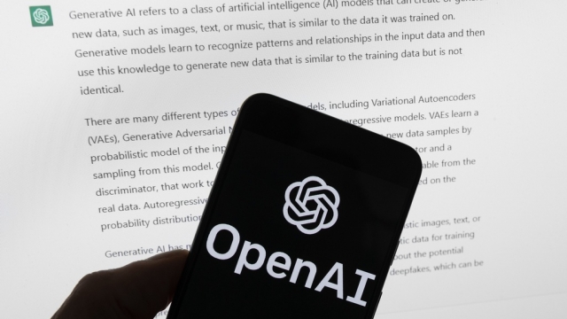 The OpenAI logo is seen on a mobile phone in front of a computer screen.