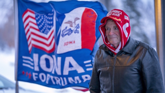 A man stands next to a flag that reads "Iowa for Trump" in Urbandale, Iowa.