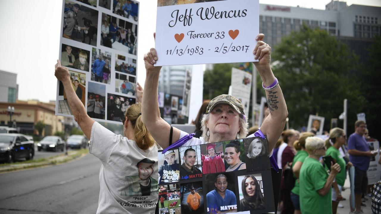 A woman holds a sign with a picture of her son and wears a sign of others' loved ones lost to OxyContin and other opioids.