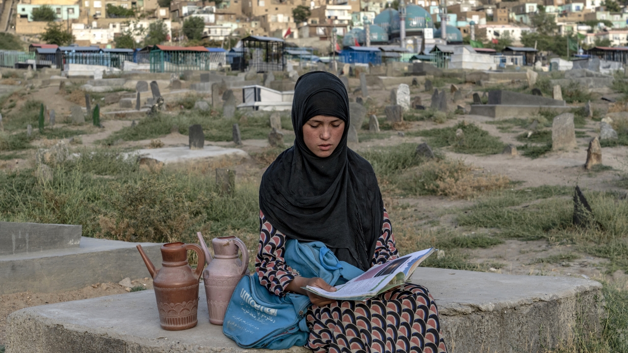 A 14-year-old Afghan girl sits on a grave and reads a book as she waits for customers to sell water at a cemetery