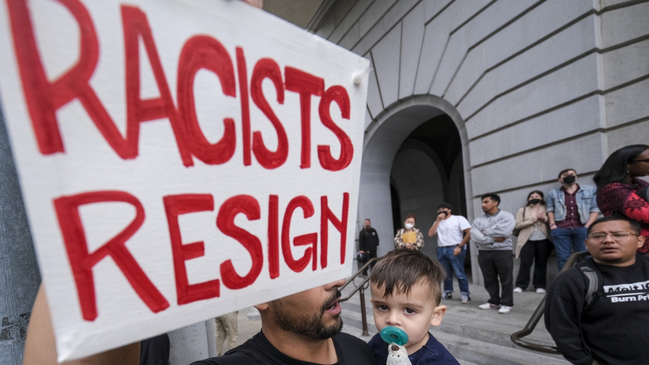 A man and his son hold a "Racists Resign" sign in protest protest outside City Hall during an LA City Council meeting.
