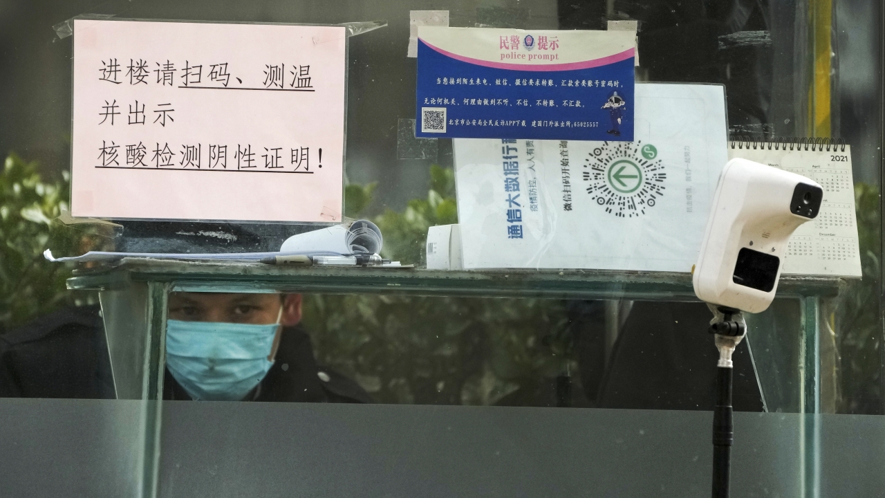 A security guard in Beijing keeps watch inside a booth displaying a temperature scan device and a travel QR code.