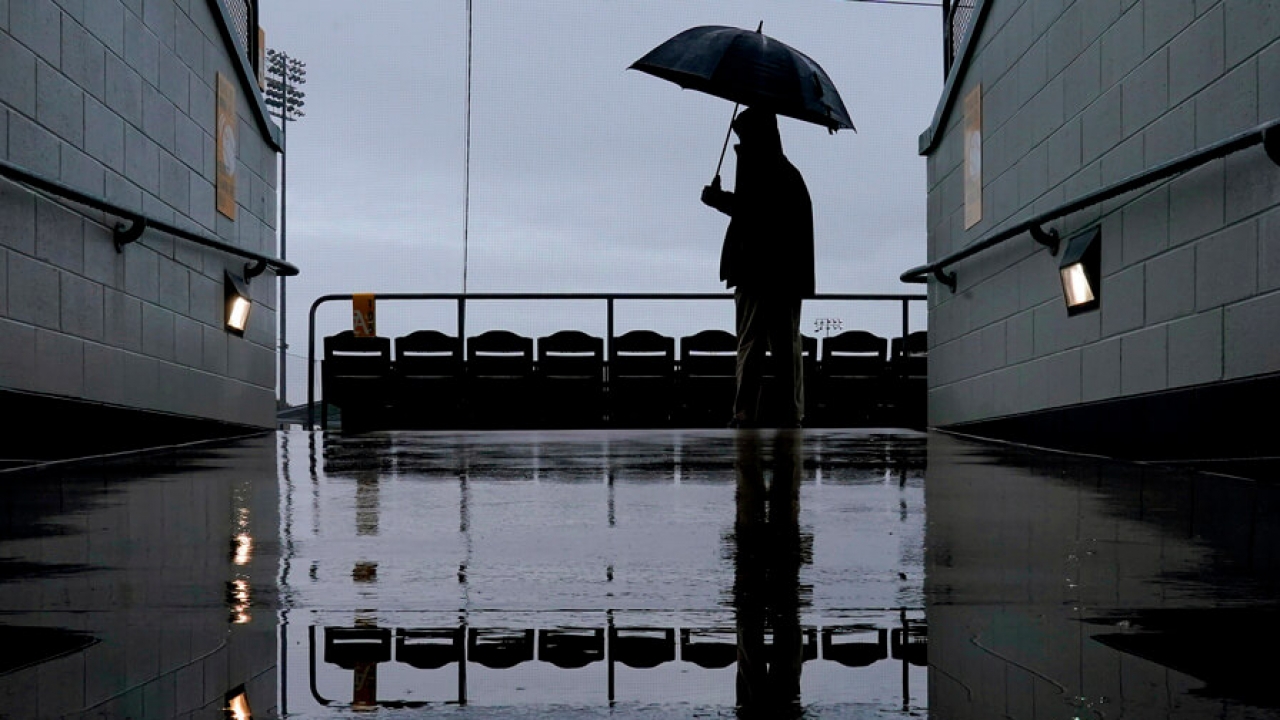 A man with an umbrella stands in the rain at a baseball stadium.