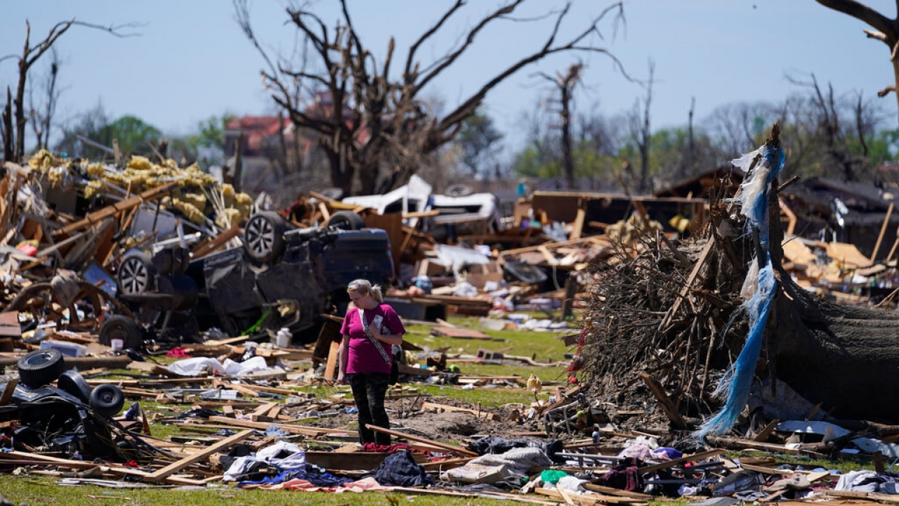 A woman walks near an uprooted tree, a flipped vehicle and debris from homes damaged by a tornado.