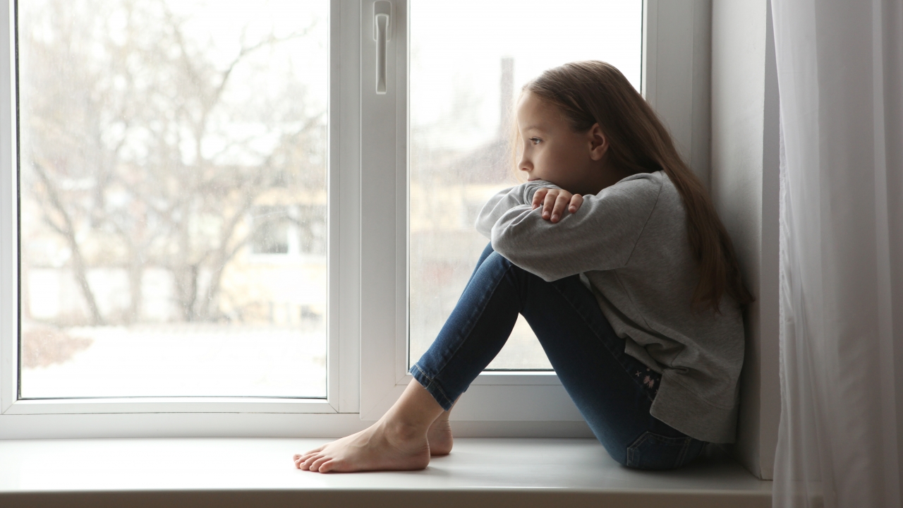 Young girl sits by window with her arms crossed over her knees