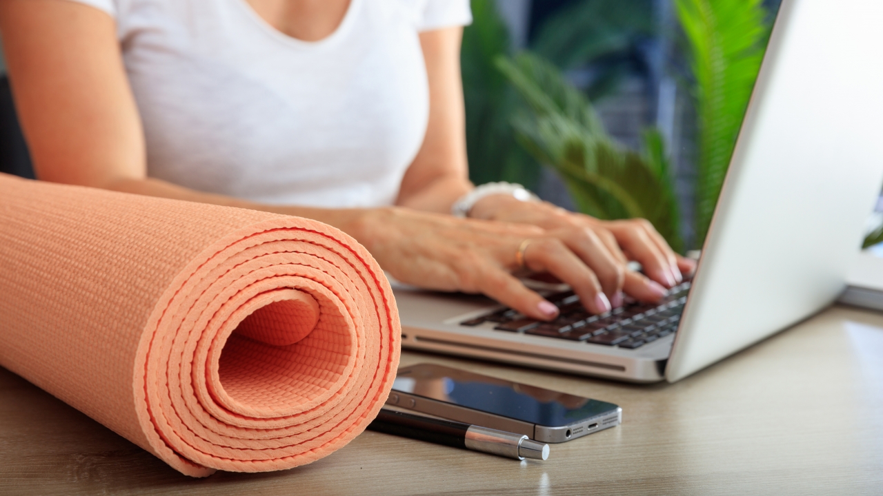 A person sits at a laptop with a yoga mat next to them.