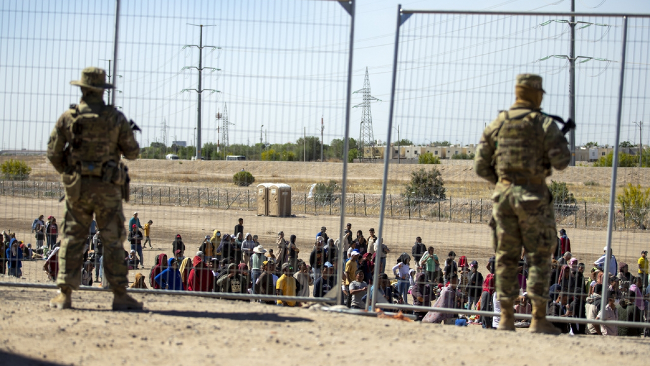 Migrants wait in line adjacent to the border fence under the watch of the Texas National Guard to enter into El Paso, Texas