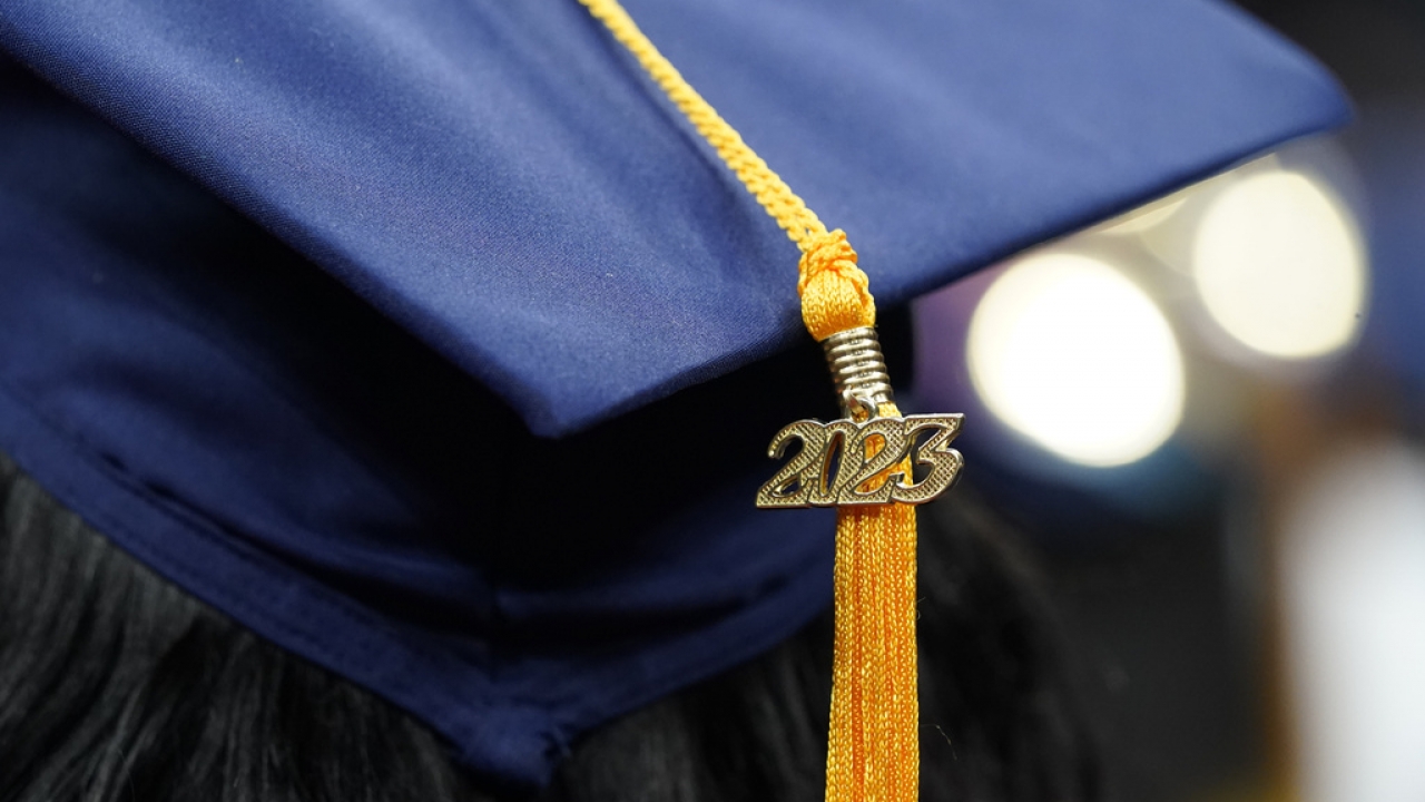 A tassel with 2023 on it rests on a graduation cap as students walk in a procession.