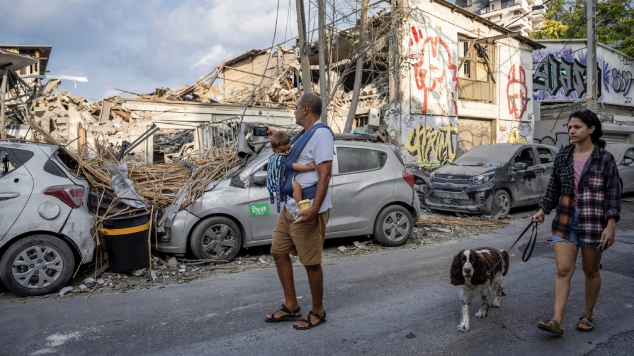 A man holding a baby while a woman walking a dog pass by rubble after Hamas attacked Israel.